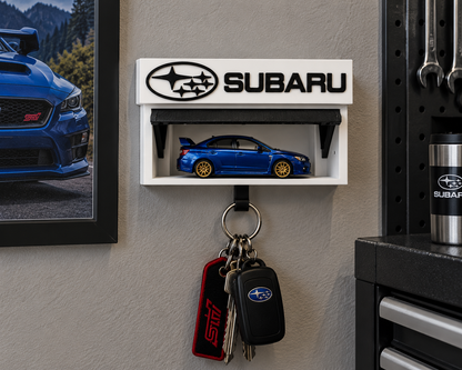 White and black Subaru key holder on a garage wall with a blue die-cast car on the shelf and keys on the hook